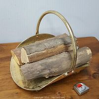 Brass firewood holder with curved handle and firewood on a wooden surface, with tape measure beside for size reference.