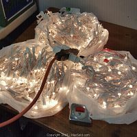 Three bundles of white Christmas lights in plastic bags, plugged in and lit, placed on a table.