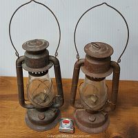 Front view of both Beacon windproof oil lamps side by side showing rust patina, clear glass globes, and metal handles. Small measuring tape placed in front for scale.