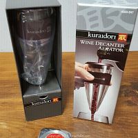 Front view of the Kuraidori wine decanter aerator in box, showing device inside plastic wrap and exterior box image illustrating usage.