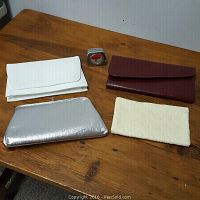 Four handbags or clutches laid out on wooden table: white, silver metallic, brown leather, cream fabric