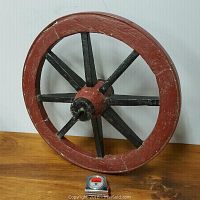 Front view of the antique wooden wheelbarrow wheel showing the reddish wooden outer rim, black spokes, steel hub, and steel rim around the edge with a measuring tape for scale.