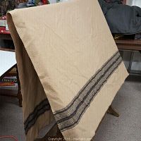 Beige wool blanket displayed folded over a stand showing black horizontal stripes near one edge. Visible texture and small mark on corner.