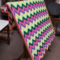 Afghan displayed over wooden stand, showing vibrant multicolored zigzag crochet pattern including pink, purple, yellow, green, and black with pink border.