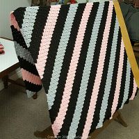Hand knit afghan draped over a wooden stand showing diagonal stripes in pink, black, and grey.