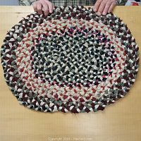 Oval braided rag rug placed flat on a wooden surface with hands holding the sides to show dimensions and texture.