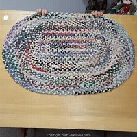 Full view of oval braided rag rug held up to show size and shape, displaying multicolored fabric strips in braided pattern.