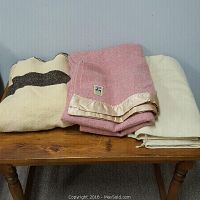 Three wool blankets folded on a wooden table - pink with satin trim, plain white, and cream with dark stripe