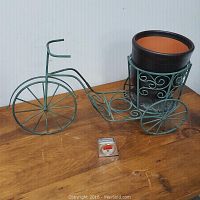 Green wire plant stand shaped like a bike with decorative scrollwork and three wheels, sitting on wooden surface next to a small measuring tape for scale.