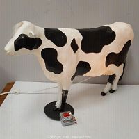 Full side view of the plastic cow sculpture showing the black and white pattern, plug-in wire, and switch on a black circular base with tape measure for scale.