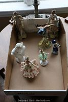 Overview of the seven small figurines placed inside a cardboard box on a table, showing various seated and standing figures including dancers and classical figures.