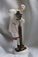 Porcelain figurine showing wigmaker standing and adjusting a wig on a stand, white colonial attire with red knee breeches and black buckle shoes.