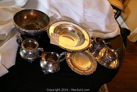 Eight-piece set of silverplated tableware displayed on black cloth with white fabric background. Shows multiple bowls, cream and sugar set, and serving tray.