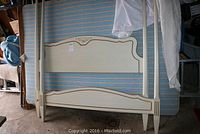 Four poster bed frame (headboard and footboard) with cream finish, decorative accents, leaning against a blue and white striped mattress with a white bedspread draped over the posts.