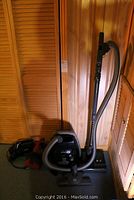 Sebo Airbelt K3 vacuum canister with extended suction wand and hose, alongside two handheld vacuums on the floor.