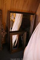 Two rectangular mirrors leaning against a wooden wall, larger mirror with brown and gold frame behind smaller ornate gold framed mirror