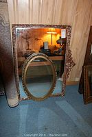Two gilt framed mirrors leaning against a wood paneled wall, the larger rectangular mirror behind an oval mirror in front.