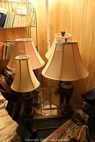 Four decorative table lamps with varying cream to beige fabric lampshades displayed on a glass surface with bookshelves in background.