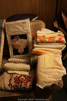 Full lot of folded tablecloths piled on a chair including one new in package and several with decorative prints.