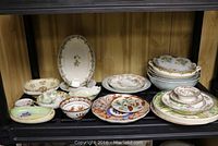 Wide view of multiple plates and bowls displayed on shelving, showing variety of patterns and sizes in the lot.