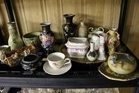 Black plastic shelf displaying multiple decorative vases, planters, cloisonné pieces, and a teacup with saucer