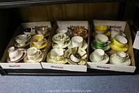 18 assorted tea cups and saucers displayed in three cardboard trays, showing a variety of floral designs and colors with gold trims