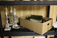 Two table lamps with clear hurricane glass shades and attached lusters sitting side by side on a shelf, next to a cardboard box containing a larger hanging lamp.