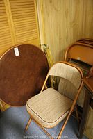 Round folding card table leaning against a wall along with folding chairs in a corner.