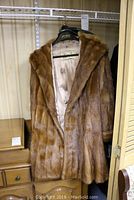 Front and side view of a brown mink fur jacket on a hanger, hung in a closet. Shows shawl collar and smooth fur texture.