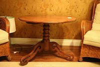 Full view of the round wooden occasional table placed between two wicker chairs, showing the entire table structure including the tabletop and legs.