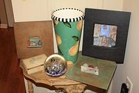 All items of the lot displayed on a wooden corner table: three framed mixed media artworks, ceramic vase, alabaster jewelry box, and blown glass paperweight