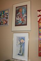 Photo showing both framed artworks hung on a wall, one smaller floral watercolor by Lyn Snow and one larger stylized horses and figures gouache by Lorene Bourgeois.