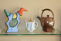 Lot image showing a ceramic bird figurine, white spouted teapot with gold accents, and brown stoneware teapot on a glass shelf against a beige wall