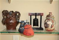 Four decorative items displayed on a glass shelf: a brown stoneware head-shaped vase, a small bull and toreador figurine, a black metal bell on a carved wooden frame with mallet, and a colorful lidded stoneware jar.