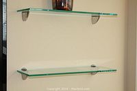 Two clear glass shelves mounted on a beige wall with brushed chrome finish brackets. Shelves are empty in this image.