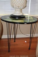 Full view of the round side table showing the laminated top with a scenic print and black hairpin metal legs on a wooden floor.