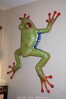 Full side profile view showing paper mache frog sculpture hanging on wall with green body, orange toes, and blue sides.