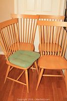 Four handmade wooden dining chairs grouped together against a wall, showing spindle backs and natural light wood finish. One chair includes a green seat cushion.