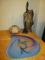 Photo showing a smooth-gradient abstract Salamandra glass art piece with curved design in front of a set of decorative items on a wooden surface.