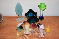 Photo showing seven colorful glass bird figurines on a branch, a green and blue vase, and a blue and black angular bowl, placed on a wooden surface.