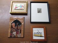 Four framed art pieces displayed on a wooden surface: enamel fruit art, cable car tile, small etching, and Jerusalem architectural tile.