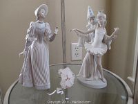 Porcelain figurines on a glass table showing lady with striped dress and parasol beside clown and ballerina figurine.