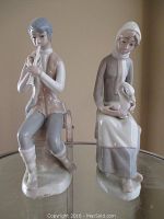 Photo showing both porcelain figurines, boy playing flute and maiden holding goose, together on glass surface
