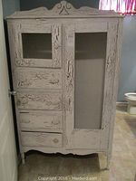Front view of distressed wood cabinet showing three drawers on left and two glass-paneled doors, with decorative carved trim on top.