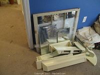 View of stacked shelving pieces, a rectangular mirror, and a large window pane decorative mirror leaning against a blue wall.