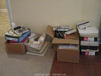 Wide view showing boxes with a receipt machine on top, stacks of paper reams, and other boxed office items on the floor.