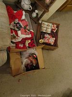 Three boxes of assorted Christmas decor including festive textiles, ceramic figurines, ornaments and plush figures arranged on carpet near furniture.