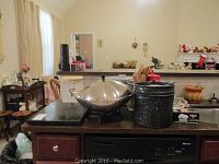 Wide view of kitchen counter showing electric wok with lid, black speckled boilers, stone board, and other items.