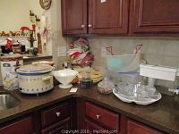 Photo showing ceramic crock pot, plastic containers, glass and ceramic serving bowls, woven mats, folded kitchen towels stacked in corner, and Brita water dispenser on countertop in kitchen.