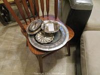 Multiple silverplate and silver-toned serving pieces including trays, bowls, and covered dishes, shown arranged on a wooden chair.